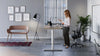 RoundE Bamboo Sit Stand Desk in White Leg Finish with Black Mesh Back Armchair and Brown Sofa in Modern Office Setting