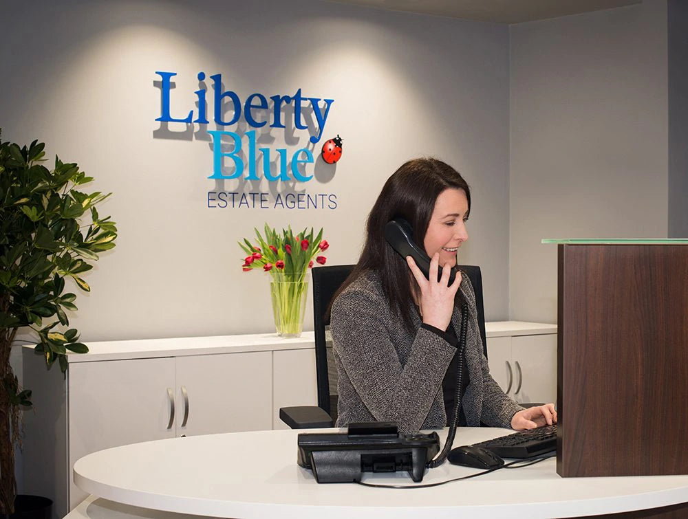 Woman In Grey Talking On Phone In Front Of Liberty Blue Logo